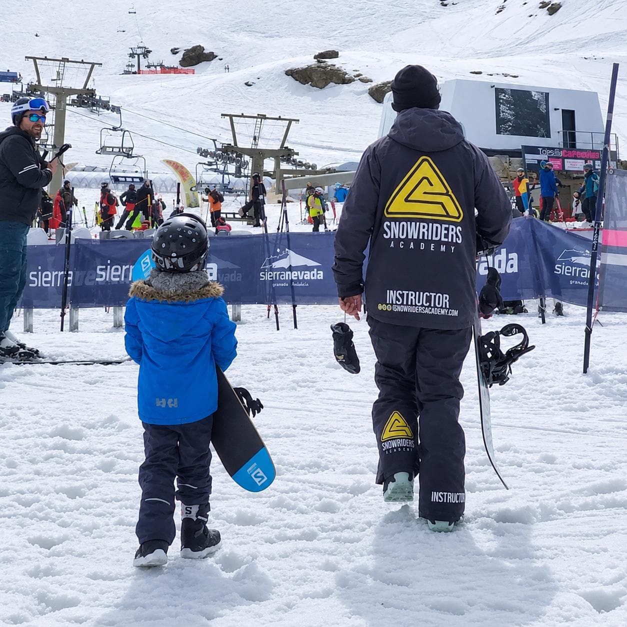 Monitor de Snow Riders Academy caminando con un niño hacia la pista, ambos con tabla de snowboard