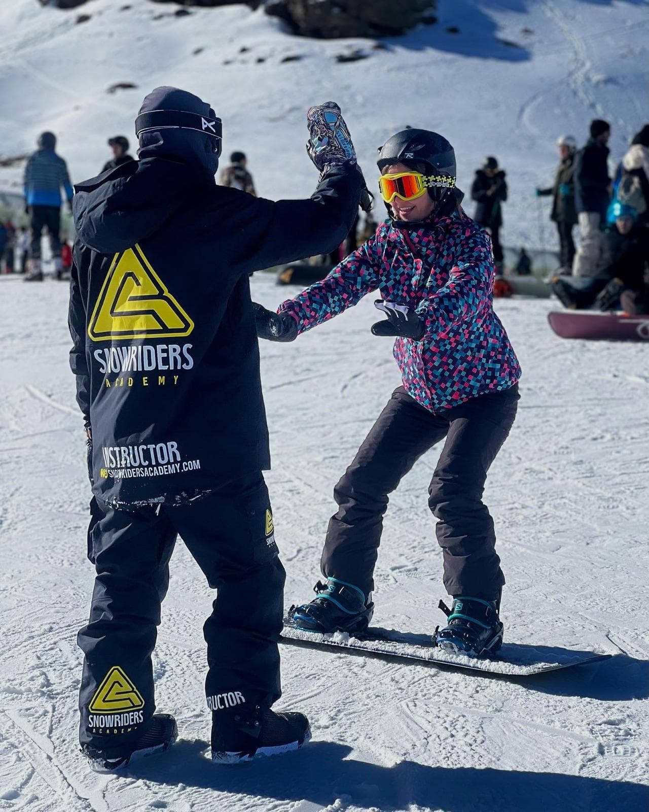 Monitor de Snow Riders Academy chocando la mano con una alumna sobre la tabla de snowboard